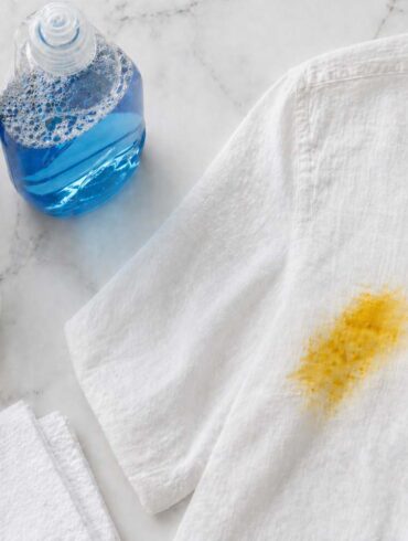 flat lay on a white marble countertop showing a glass bowl with baking soda paste, a small bottle of hydrogen peroxide, a bottle of dish soap, a clean white cloth folded neatly, and a white linen shirt with a turmeric curry stain visible