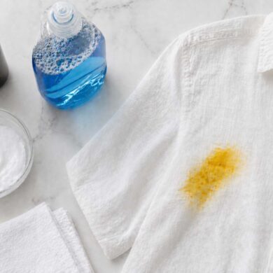 flat lay on a white marble countertop showing a glass bowl with baking soda paste, a small bottle of hydrogen peroxide, a bottle of dish soap, a clean white cloth folded neatly, and a white linen shirt with a turmeric curry stain visible