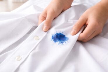 Close-up of two hands holding a crisp white button-down shirt, gently pulling the fabric taut to reveal a vivid blue ink stain near the front placket. The stain has a darker center with softer, feathered edges, suggesting it has soaked into the fabric. The background is softly blurred, with bright, natural light highlighting the texture of the cloth and the contrast between the clean white shirt and the stain.