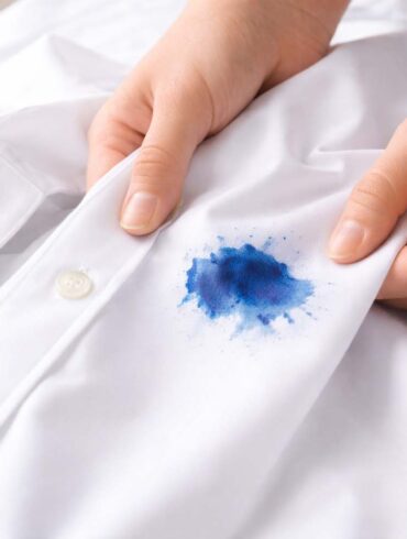 Close-up of two hands holding a crisp white button-down shirt, gently pulling the fabric taut to reveal a vivid blue ink stain near the front placket. The stain has a darker center with softer, feathered edges, suggesting it has soaked into the fabric. The background is softly blurred, with bright, natural light highlighting the texture of the cloth and the contrast between the clean white shirt and the stain.