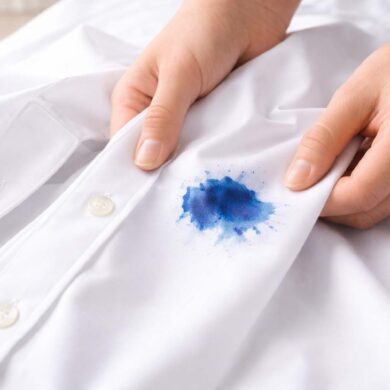 Close-up of two hands holding a crisp white button-down shirt, gently pulling the fabric taut to reveal a vivid blue ink stain near the front placket. The stain has a darker center with softer, feathered edges, suggesting it has soaked into the fabric. The background is softly blurred, with bright, natural light highlighting the texture of the cloth and the contrast between the clean white shirt and the stain.