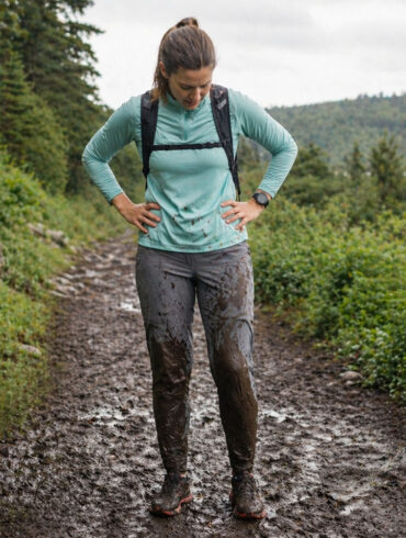 A woman standing in a muddy trail running setting, looking down at brown mud splattered up both shins and across the front of gray running pants, expression resigned. Lush green trail visible behind them, overcast sky. The mud is clearly visible and dramatic
