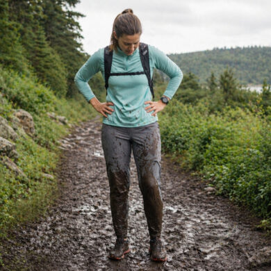 A woman standing in a muddy trail running setting, looking down at brown mud splattered up both shins and across the front of gray running pants, expression resigned. Lush green trail visible behind them, overcast sky. The mud is clearly visible and dramatic