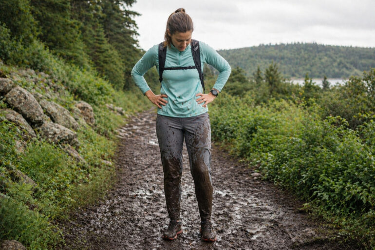 A woman standing in a muddy trail running setting, looking down at brown mud splattered up both shins and across the front of gray running pants, expression resigned. Lush green trail visible behind them, overcast sky. The mud is clearly visible and dramatic