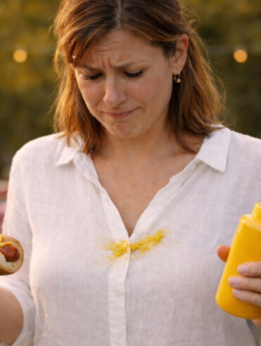A woman in her late 30s at a summer backyard cookout, looking down at a bright yellow mustard splatter across the front of a white linen shirt with a mix of disbelief and resignation. She's holding a half-eaten hot dog in a bun in one hand, a mustard squeeze bottle in the other, the classic squeeze-too-hard moment written all over her face. Picnic table behind her with paper plates, condiments, and a cooler visible