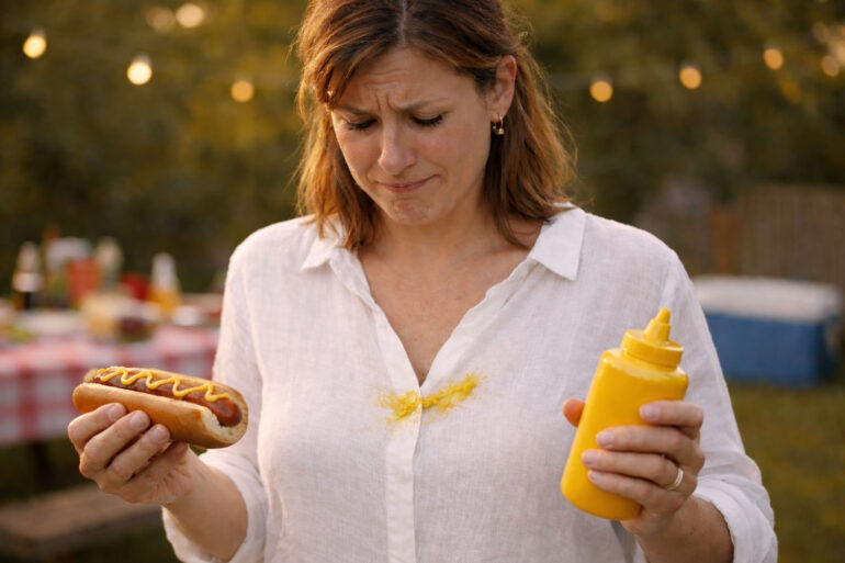 A woman in her late 30s at a summer backyard cookout, looking down at a bright yellow mustard splatter across the front of a white linen shirt with a mix of disbelief and resignation. She's holding a half-eaten hot dog in a bun in one hand, a mustard squeeze bottle in the other, the classic squeeze-too-hard moment written all over her face. Picnic table behind her with paper plates, condiments, and a cooler visible