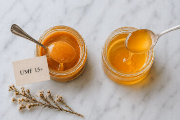 Overhead view of two small glass jars of honey on a pale marble surface. The left jar, labeled “UMF 15+,” contains thick, amber-colored honey with a smooth, glossy texture; a spoon lifts a rounded scoop with a slow drip falling back into the jar. The right jar holds a lighter, more translucent honey with a thinner consistency, also shown dripping from a spoon. A small sprig of delicate white flowers rests nearby, adding a soft decorative touch.