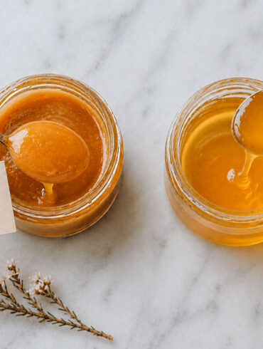 Overhead view of two small glass jars of honey on a pale marble surface. The left jar, labeled “UMF 15+,” contains thick, amber-colored honey with a smooth, glossy texture; a spoon lifts a rounded scoop with a slow drip falling back into the jar. The right jar holds a lighter, more translucent honey with a thinner consistency, also shown dripping from a spoon. A small sprig of delicate white flowers rests nearby, adding a soft decorative touch.