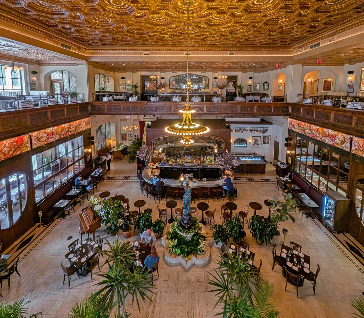 The soaring interior of La Grande Boucherie DC restaurant photographed from the mezzanine level, showing the spectacular gilded coffered ceiling with intricate gold geometric patterns, a grand brass and glass chandelier, Art Nouveau stained glass panels in warm reds and oranges along the mezzanine balustrade, a central circular bar with dark wood surround, tropical potted plants throughout the main floor, and diners seated at bistro tables below