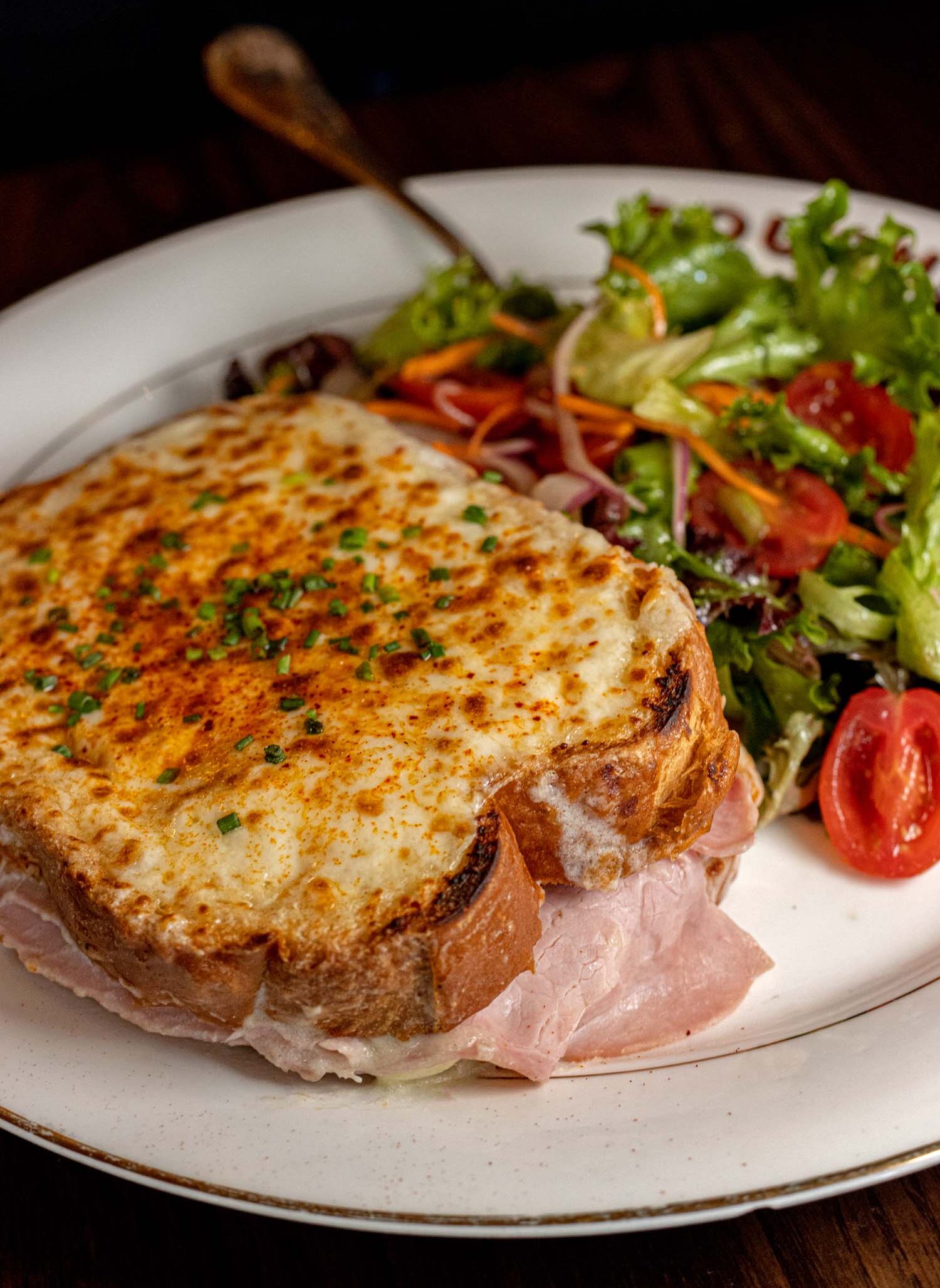 A classic Croque Monsieur served at La Grande Boucherie DC, presented on a round white plate with a gold rim, showing thick-cut brioche toast topped with a deeply golden broiled Gruyère crust sprinkled with chopped chives, with slices of jambon ham visible spilling from the sides of the sandwich, accompanied by a bright fresh petite salad of mixed greens, cherry tomatoes, shredded carrot, and red onion, with a vintage brass fork resting in the background against a dark wood surface