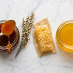 Overhead flat lay on honed white marble. Two open jars of honey side by side, one visibly darker and thicker (manuka), one lighter and more fluid (raw wildflower). A single silver spoon rests across the manuka jar with a slow drip of dark amber honey. Between the jars, a small sprig of dried manuka flowers and a single raw honeycomb fragment on a piece of natural parchment.