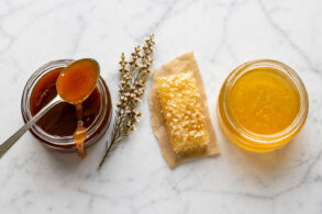 Overhead flat lay on honed white marble. Two open jars of honey side by side, one visibly darker and thicker (manuka), one lighter and more fluid (raw wildflower). A single silver spoon rests across the manuka jar with a slow drip of dark amber honey. Between the jars, a small sprig of dried manuka flowers and a single raw honeycomb fragment on a piece of natural parchment.