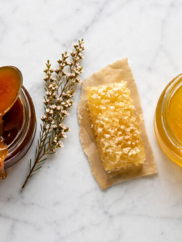 Overhead flat lay on honed white marble. Two open jars of honey side by side, one visibly darker and thicker (manuka), one lighter and more fluid (raw wildflower). A single silver spoon rests across the manuka jar with a slow drip of dark amber honey. Between the jars, a small sprig of dried manuka flowers and a single raw honeycomb fragment on a piece of natural parchment.