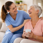 A smiling caregiver in blue scrubs leans in and gently holds the shoulders of an elderly woman seated on a couch. The older woman, with short gray hair, smiles back warmly while holding a white mug. They make eye contact, sharing a moment of connection in a bright, cozy living room with soft natural light, a bookshelf, and a sofa in the background.