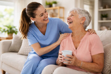 A smiling caregiver in blue scrubs leans in and gently holds the shoulders of an elderly woman seated on a couch. The older woman, with short gray hair, smiles back warmly while holding a white mug. They make eye contact, sharing a moment of connection in a bright, cozy living room with soft natural light, a bookshelf, and a sofa in the background.