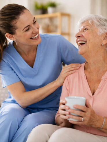 A smiling caregiver in blue scrubs leans in and gently holds the shoulders of an elderly woman seated on a couch. The older woman, with short gray hair, smiles back warmly while holding a white mug. They make eye contact, sharing a moment of connection in a bright, cozy living room with soft natural light, a bookshelf, and a sofa in the background.