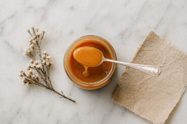 Overhead flat lay on honed white marble. A single open jar of very manuka honey centered in frame. A silver spoon rests across the jar with a slow honey drip. Beside it, a small dried manuka flower sprig and a folded piece of natural parchment.