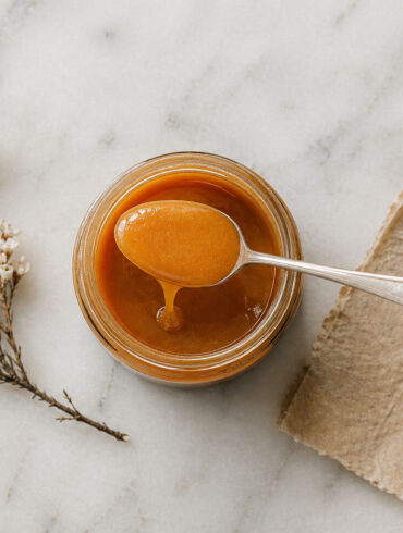 Overhead flat lay on honed white marble. A single open jar of very manuka honey centered in frame. A silver spoon rests across the jar with a slow honey drip. Beside it, a small dried manuka flower sprig and a folded piece of natural parchment.