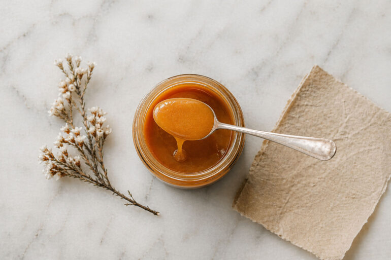 Overhead flat lay on honed white marble. A single open jar of very manuka honey centered in frame. A silver spoon rests across the jar with a slow honey drip. Beside it, a small dried manuka flower sprig and a folded piece of natural parchment.