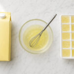 Clean flat lay on white marble. Center: a glass bowl of clear egg whites with a small whisk. Left: a sealed carton of liquid egg whites. Right: an ice cube tray with frozen egg white portions.