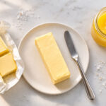one unwrapped stick of butter on a small white ceramic plate, a clean butter knife resting beside it. Left: a halved stick still in its gold foil wrapper showing the date stamp end. Right: a small glass jar of golden ghee with the lid off.
