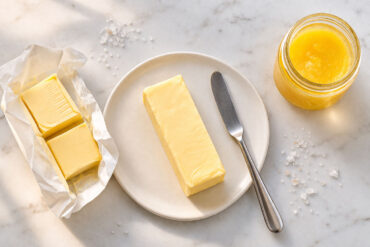 one unwrapped stick of butter on a small white ceramic plate, a clean butter knife resting beside it. Left: a halved stick still in its gold foil wrapper showing the date stamp end. Right: a small glass jar of golden ghee with the lid off.