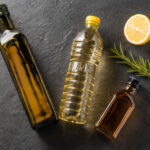 Three different bottles of cooking oil arranged loosely: one olive oil (dark glass), one clear vegetable oil bottle, one small amber bottle. A halved lemon and a sprig of fresh rosemary nearby.