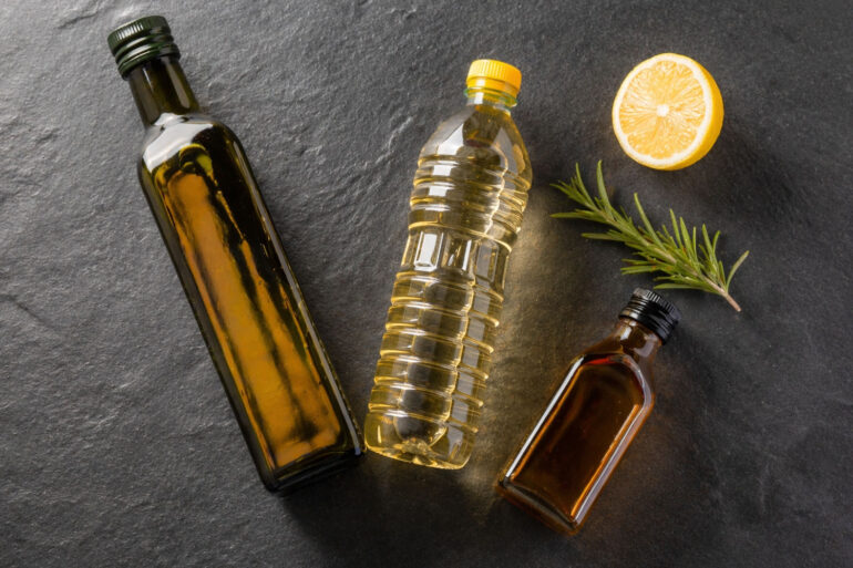 Three different bottles of cooking oil arranged loosely: one olive oil (dark glass), one clear vegetable oil bottle, one small amber bottle. A halved lemon and a sprig of fresh rosemary nearby.