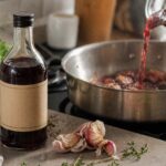 Bottle of cooking wine in the foreground as red wine is poured into a skillet with garlic cloves and fresh herbs in a softly lit kitchen