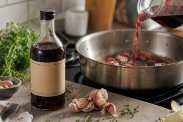 Bottle of cooking wine in the foreground as red wine is poured into a skillet with garlic cloves and fresh herbs in a softly lit kitchen