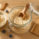 an open glass canister of rolled oats with a wooden scoop. Left: a small bowl with a cooked portion of oatmeal. Right: a single flavored instant oatmeal packet. Scattered props: a few blueberries and a cinnamon stick