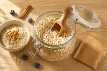 an open glass canister of rolled oats with a wooden scoop. Left: a small bowl with a cooked portion of oatmeal. Right: a single flavored instant oatmeal packet. Scattered props: a few blueberries and a cinnamon stick