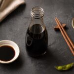 Flat lay on a dark slate surface. Center: a dark glass bottle of soy sauce with the cap off. Left: a small ceramic dipping dish with soy sauce. Right: a pair of chopsticks and a few edamame pods.