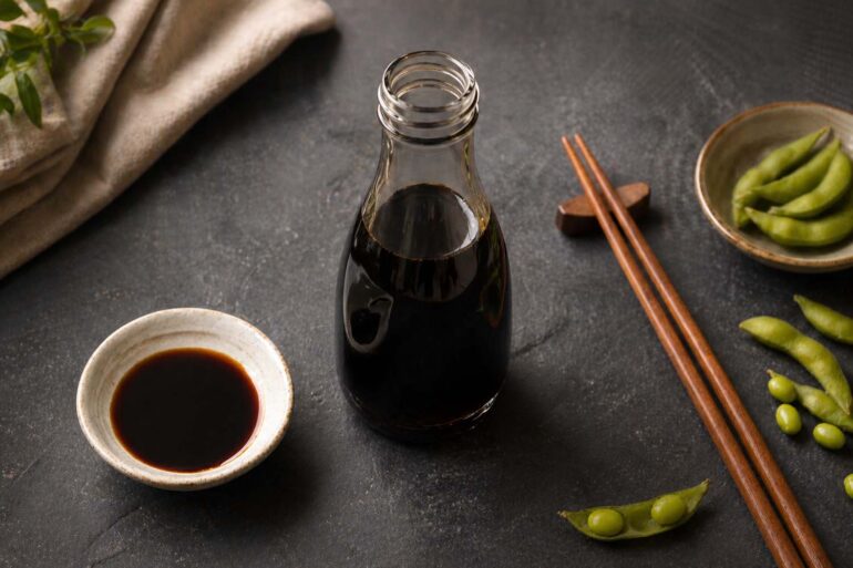 Flat lay on a dark slate surface. Center: a dark glass bottle of soy sauce with the cap off. Left: a small ceramic dipping dish with soy sauce. Right: a pair of chopsticks and a few edamame pods.