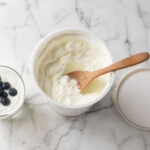 Flat lay on white marble. Center: an open tub of Greek yogurt with visible whey pooling on top and a clean wooden spoon. Left: a small glass of plain yogurt with fresh blueberries. Right: the container lid