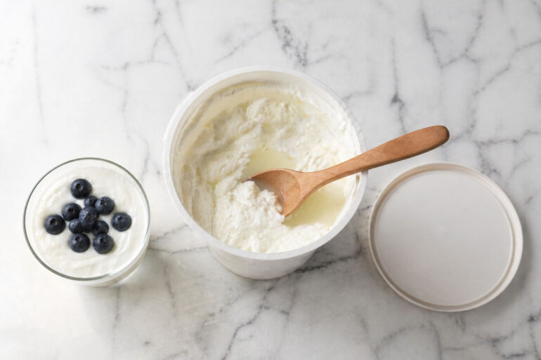 Flat lay on white marble. Center: an open tub of Greek yogurt with visible whey pooling on top and a clean wooden spoon. Left: a small glass of plain yogurt with fresh blueberries. Right: the container lid