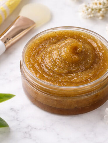 Overhead photo of a small glass jar filled with manuka honey swirled into a soft peak. The jar sits on a white marble surface. Surrounding it are a folded yellow-and-white striped towel, a white-handled cosmetic spatula with a gold ferrule, small white flowers, and a few green leaves. Soft natural light creates gentle shadows, giving the scene a clean, spa-like feel.