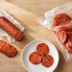 a whole pepperoni stick, partially unwrapped, with a few slices cut and fanned out beside it. Right side: an open vacuum-sealed bag of sliced pepperoni. Center bottom: a small white plate with three or four pepperoni slices laid flat