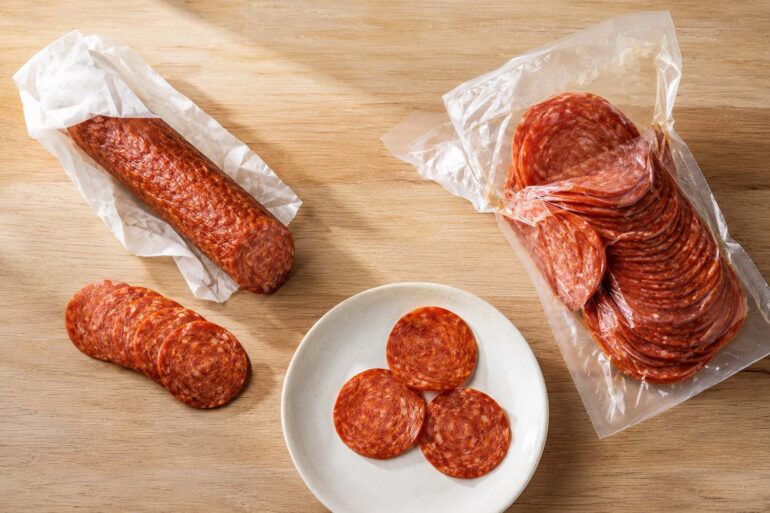 a whole pepperoni stick, partially unwrapped, with a few slices cut and fanned out beside it. Right side: an open vacuum-sealed bag of sliced pepperoni. Center bottom: a small white plate with three or four pepperoni slices laid flat