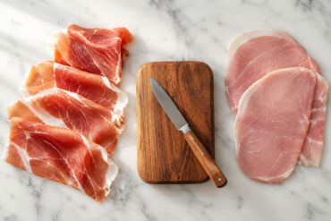 Overhead flat lay on a white marble surface. Left side: three or four paper-thin slices of prosciutto crudo draped loosely, showing the translucent ruby-red meat and white fat ribbons. Right side: two or three thicker, pale pink slices of prosciutto cotto laid flat for contrast. Center: a small wooden board with a knife resting at an angle.
