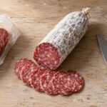 Overhead flat lay on a worn light wood surface. Center: a whole dry-cured salami, sliced open to reveal the interior, with four or five slices fanned out showing the red-pink interior and white fat marbling. The cut end clearly shows the white mold casing. Left: a small open package of sliced deli salami. Right: a knife resting at an angle.