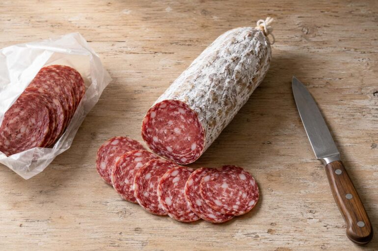 Overhead flat lay on a worn light wood surface. Center: a whole dry-cured salami, sliced open to reveal the interior, with four or five slices fanned out showing the red-pink interior and white fat marbling. The cut end clearly shows the white mold casing. Left: a small open package of sliced deli salami. Right: a knife resting at an angle.