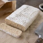 A rectangular block of tempeh on a worn light wood cutting board, visible white mycelium coating on the surface with small natural black spore spots scattered across the top. One clean slice cut from the block showing the beige interior with soybeans bound together. A chef's knife resting beside it