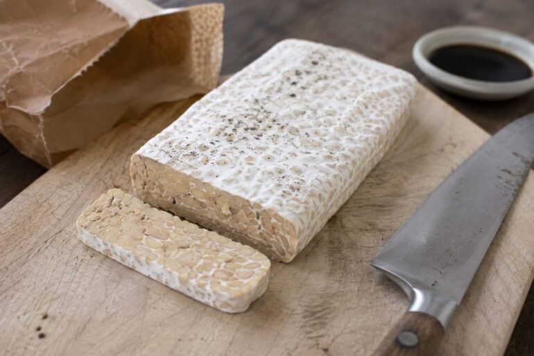 A rectangular block of tempeh on a worn light wood cutting board, visible white mycelium coating on the surface with small natural black spore spots scattered across the top. One clean slice cut from the block showing the beige interior with soybeans bound together. A chef's knife resting beside it