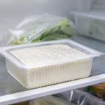 Unopened water-packed tofu tub sitting on a refrigerator shelf, cold white interior light, slight condensation on the plastic, other fridge items softly blurred in background