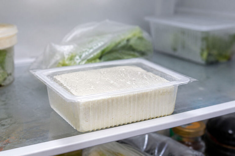 Unopened water-packed tofu tub sitting on a refrigerator shelf, cold white interior light, slight condensation on the plastic, other fridge items softly blurred in background