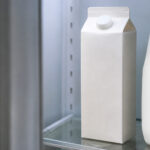 Carton of half and half on a refrigerator shelf, upright and sealed. Cool fridge interior light. Partial view of a milk jug alongside it. Door slightly open