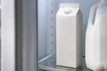 Carton of half and half on a refrigerator shelf, upright and sealed. Cool fridge interior light. Partial view of a milk jug alongside it. Door slightly open