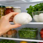 Hand holding a wedge of mozzarella cheese inside a clean, well-lit refrigerator, with neatly arranged jars, eggs, greens, and vegetables on white shelves in the background.