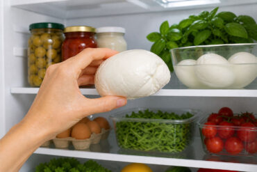 Hand holding a wedge of mozzarella cheese inside a clean, well-lit refrigerator, with neatly arranged jars, eggs, greens, and vegetables on white shelves in the background.
